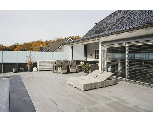 Moderne Terrasse mit hellgrauen Bodenplatten, hellen Rattan-Gartenmöbeln, Sonnenliegen und Sichtschutz aus Glas.