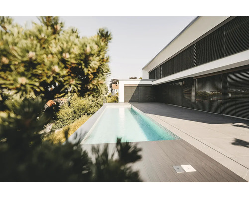 Modernes weißes Wohnhaus mit schwarzen Außenjalousien, einem schmalen Swimmingpool und einer großzügigen Terrasse mit grauen Steinplatten.