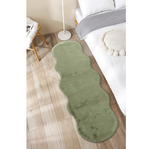 Grüner Kunstfell-Läufer mit gewellten Kanten in einem hellen Schlafzimmer auf Holzboden neben einem Bett.