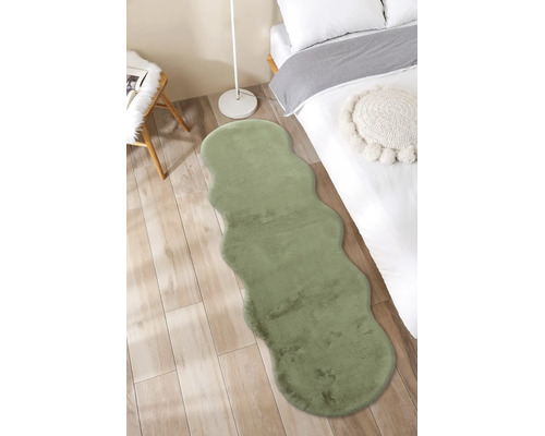 Grüner Kunstfell-Läufer mit gewellten Kanten in einem hellen Schlafzimmer auf Holzboden neben einem Bett.
