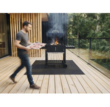 Ein Mann trägt ein Tablett mit Fleisch zu einem rauchenden Holzkohle-Wagengrill auf einer Holzterrasse mit Glasgeländer und Grillmatte.