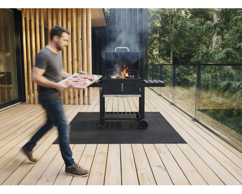Ein Mann trägt ein Tablett mit Fleisch zu einem rauchenden Holzkohle-Wagengrill auf einer Holzterrasse mit Glasgeländer und Grillmatte.
