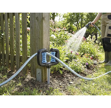 Guide-tuyau d angle pour jardin, fixé à un poteau de clôture en bois, avec un tuyau d arrosage et une personne arrosant en arrière-plan.