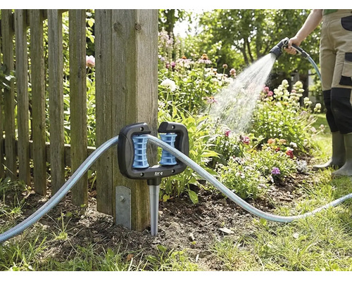 Guide-tuyau d angle pour jardin, fixé à un poteau de clôture en bois, avec un tuyau d arrosage et une personne arrosant en arrière-plan.