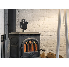 Poêle à bois noir en fonte avec feu allumé, ventilateur de poêle sur le dessus et serviteur de cheminée devant un mur en briques blanches.