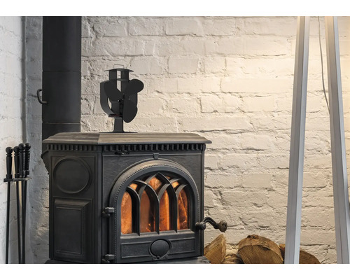 Poêle à bois noir en fonte avec feu allumé, ventilateur de poêle sur le dessus et serviteur de cheminée devant un mur en briques blanches.