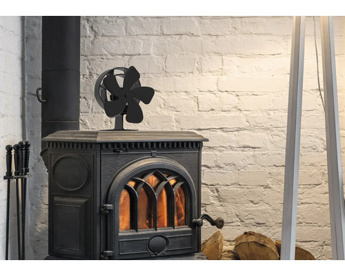 Un ventilateur de poêle noir à cinq pales sur un poêle à bois en fonte allumé devant un mur de briques blanches avec du bois de chauffage.