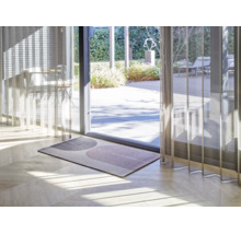 Paillasson moderne avec motif géométrique en arche sur parquet clair devant une porte vitrée de terrasse avec rideaux transparents.