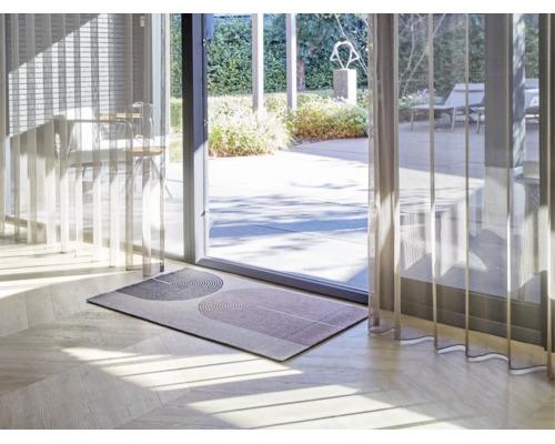 Paillasson moderne avec motif géométrique en arche sur parquet clair devant une porte vitrée de terrasse avec rideaux transparents.