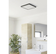 Salle de bains moderne avec plafonnier carré, meuble de rangement étroit aspect bois, lavabo blanc et plante verte.