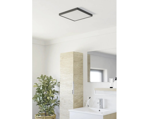 Salle de bains moderne avec plafonnier carré, meuble de rangement étroit aspect bois, lavabo blanc et plante verte.