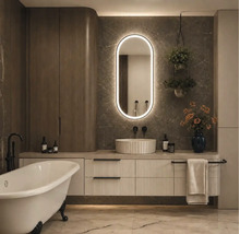 Modernes Badezimmer mit freistehender Badewanne, Unterschrank mit Rillenfront, Aufsatzwaschbecken und beleuchtetem Ovalspiegel vor Marmorwänden.