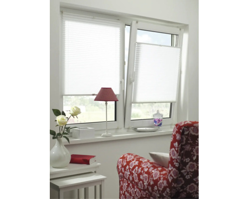 Store plissé blanc sur une fenêtre à deux vantaux dans une pièce lumineuse avec fauteuil rouge, petite table et fleurs.