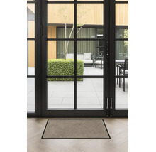 Paillasson rectangulaire avec bordure foncée sur un parquet clair devant une porte-fenêtre vitrée donnant sur une terrasse.