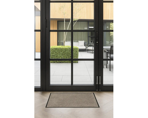 Paillasson rectangulaire avec bordure foncée sur un parquet clair devant une porte-fenêtre vitrée donnant sur une terrasse.