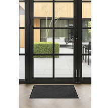 Paillasson gris foncé devant une porte vitrée noire sur un parquet clair. Vue sur une terrasse lumineuse et moderne.