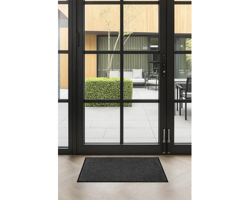 Paillasson gris foncé devant une porte vitrée noire sur un parquet clair. Vue sur une terrasse lumineuse et moderne.