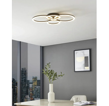 Plafonnier LED moderne avec quatre anneaux lumineux superposés dans une pièce lumineuse aux murs gris, table avec vase et vue sur la ville.