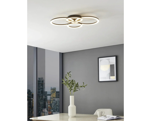Plafonnier LED moderne avec quatre anneaux lumineux superposés dans une pièce lumineuse aux murs gris, table avec vase et vue sur la ville.