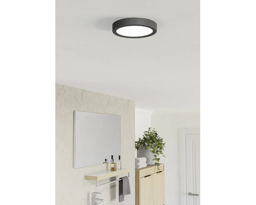 Plafonnier LED rond avec cadre sombre dans une salle de bains moderne et lumineuse avec miroir, plante verte et meuble en bois.