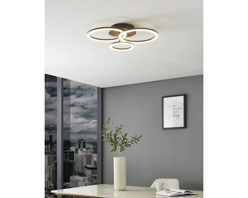 Plafonnier LED moderne à quatre anneaux lumineux dans une pièce lumineuse avec mur gris, vue sur la ville et table blanche.