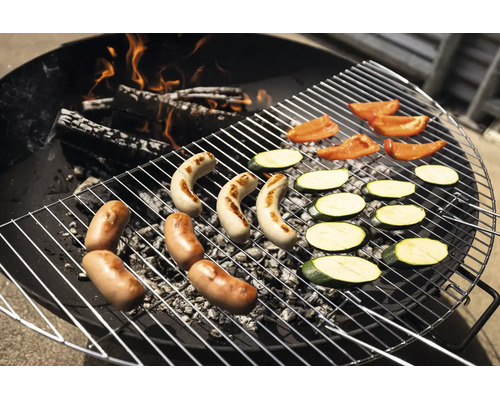 Une grille de barbecue ronde en métal sur du charbon de bois et des bûches en feu, avec des saucisses, des tranches de courgettes et des poivrons.