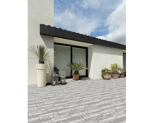 Terrasse lumineuse avec pavés gris rectangulaires modernes, décorée de bacs à plantes et de lanternes contre une façade blanche.