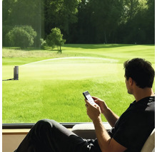 Mann steuert einen smarten Gartenregner per Smartphone-App mit Blick auf eine grosse Rasenfläche.