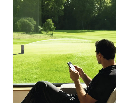Mann steuert einen smarten Gartenregner per Smartphone-App mit Blick auf eine grosse Rasenfläche.