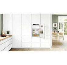 Réfrigérateur encastrable dans une cuisine moderne blanche avec porte ouverte, intérieur avec étagères en verre et tiroir à légumes, design épuré.