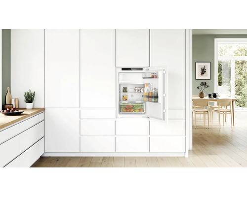 Réfrigérateur encastrable dans une cuisine moderne blanche avec porte ouverte, intérieur avec étagères en verre et tiroir à légumes, design épuré.