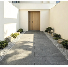 Moderner Eingangsbereich mit einem Gehweg aus grossen grauen Terrassenplatten, gesäumt von weissem Kies, kleinen Sträuchern und Steinen.