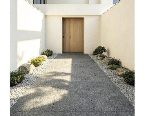Moderner Eingangsbereich mit einem Gehweg aus grossen grauen Terrassenplatten, gesäumt von weissem Kies, kleinen Sträuchern und Steinen.