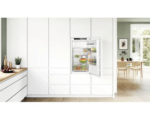 Réfrigérateur encastrable dans une cuisine blanche moderne avec porte ouverte et aliments, dans une ambiance lumineuse et chaleureuse.