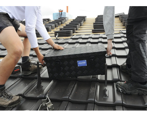 Zwei Handwerker tragen eine schwarze Aluminium Werkzeugkiste von Alutec auf einem mit Dachziegeln gedeckten Hausdach.
