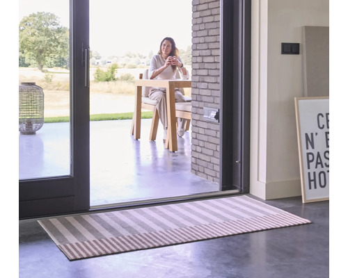Un tapis couloir rayé devant une grande porte-fenêtre donnant sur une femme assise à une table de jardin.