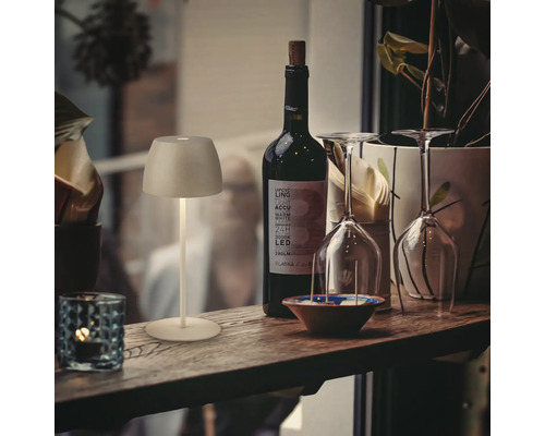 Une petite lampe LED sans fil rechargeable sur un rebord de fenêtre à côté d une bouteille de vin, de verres et d une plante, éclairage d ambiance.