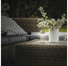 Une petite lampe LED sans fil est posée sur une table de jardin en osier à côté d'un vase avec des fleurs blanches sur une terrasse.