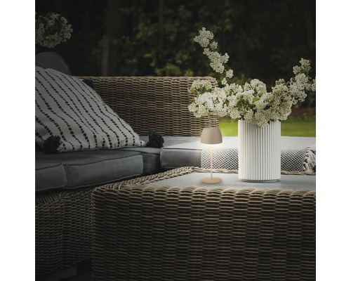Une petite lampe LED sans fil est posée sur une table de jardin en osier à côté d'un vase avec des fleurs blanches sur une terrasse.