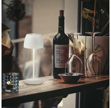 Moderne LED Akkuleuchte als Tischlampe auf einem Holztisch neben einer Weinflasche und Gläsern für eine stimmungsvolle Beleuchtung.