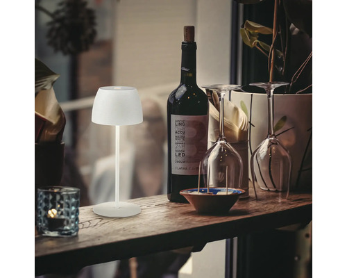 Moderne LED Akkuleuchte als Tischlampe auf einem Holztisch neben einer Weinflasche und Gläsern für eine stimmungsvolle Beleuchtung.