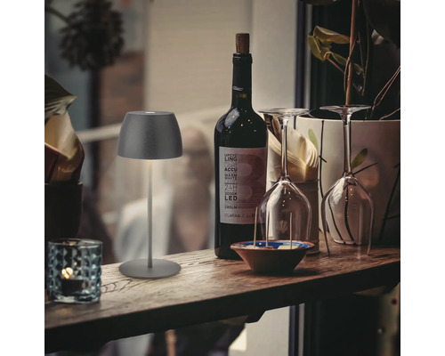 Moderne LED Tischleuchte fuer den Innen und Aussenbereich platziert auf einer rustikalen Holztheke neben einer Flasche Wein und Weinglaesern.