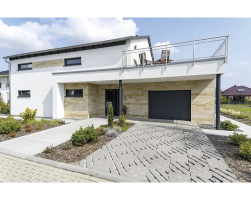 Modernes Einfamilienhaus mit Natursteinverkleidung, Garage, Balkon und einer Pflasterung aus Rasengittersteinen im Vorgarten.