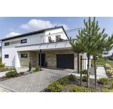 Modernes Wohnhaus mit Natursteinverkleidung am Erdgeschoss, Garage, Balkon mit Edelstahlgeländer und gepflasterter Zufahrt in einem Garten.