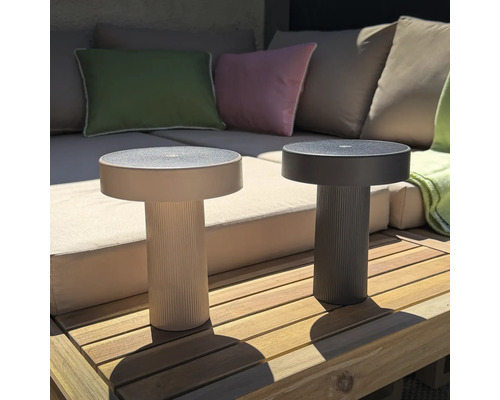 Zwei runde, moderne LED Tischleuchten mit geriffeltem Standfuß auf einer Holzterrasse vor einem Lounge Sofa.