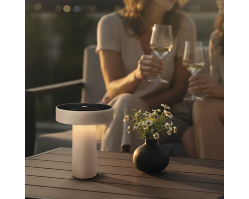 Moderne Solar Tischleuchte mit integriertem Solarpanel auf einem Terrassentisch neben einer Blumenvase bei zwei Personen mit Weingläsern.