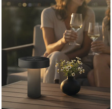 Moderne LED Tischleuchte fuer den Aussenbereich mit Solarpanel auf einem Gartentisch neben einer Blumenvase und Personen im Hintergrund