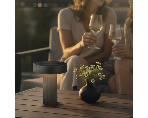 Moderne LED Tischleuchte fuer den Aussenbereich mit Solarpanel auf einem Gartentisch neben einer Blumenvase und Personen im Hintergrund