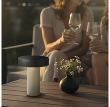 Moderne LED Tischleuchte fuer den Aussenbereich auf einem Terrassentisch neben einer Blumenvase mit zwei Personen im Hintergrund