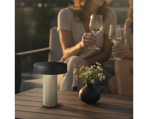 Moderne LED Tischleuchte fuer den Aussenbereich auf einem Terrassentisch neben einer Blumenvase mit zwei Personen im Hintergrund